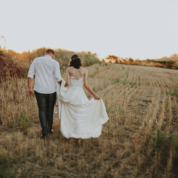 Gartenhochzeit Boho Vibes Heiraten im Grünen Sommerhochzeit Ein Paar in Hochzeitskleidung geht Hand in Hand durch ein Feld.
