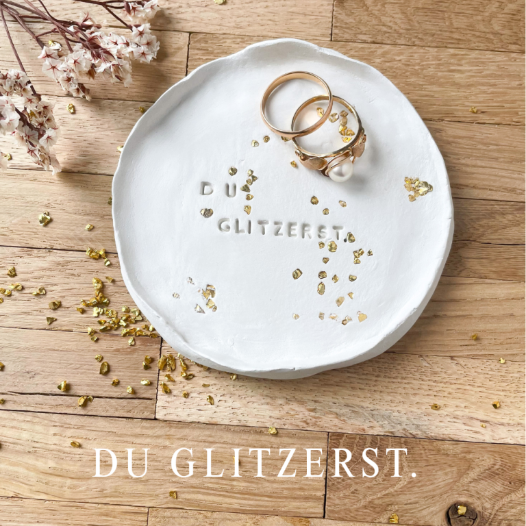 Weiße Schmuckschale mit goldenen Ringen und Glitzer, neben einer Blütenzweig.